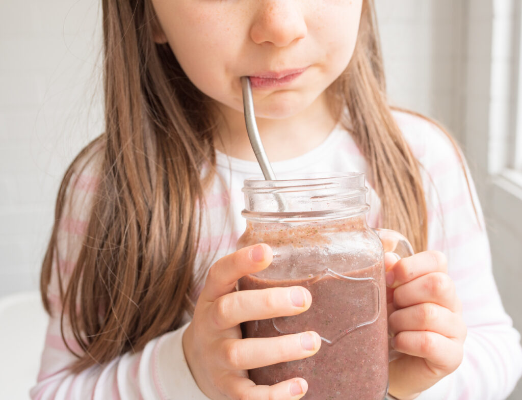 Cropped close up of little girl drinking berry smoothie from glass mug with metal straw (selective focus)